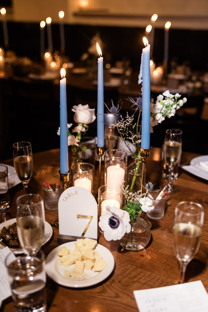 A candlelit guest table setting at Osteria Via Stato features blue taper candles, flowers, cheese, and a table marker