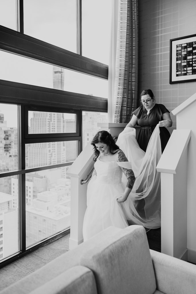 A bride descends stairs as another woman helps with her dress in a modern, city-view room, preparing for a romantic celebration at Osteria Via Stato.