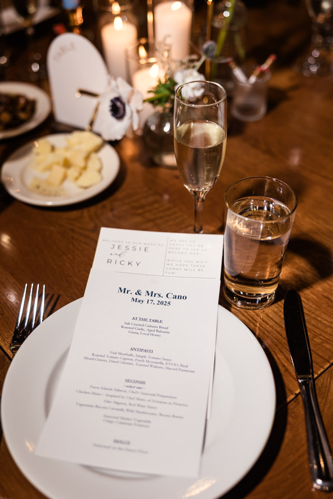 Guest place setting for wedding reception at Osteria Via Stato, featuring a menu, champagne, water, candles, and flowers on a wooden table