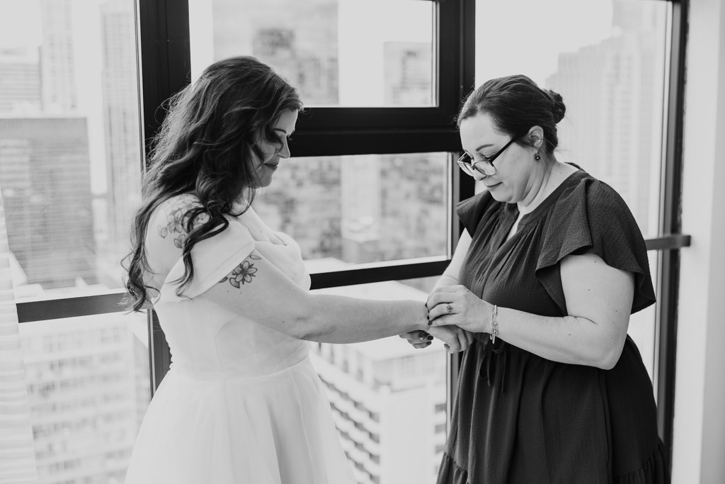 Two women stand by a window at Osteria Via Stato; one helps the bride with her bracelet in a bright, city-view room