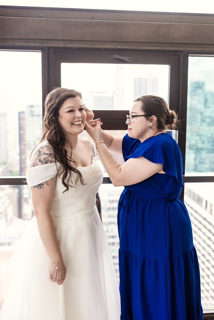 Bridesmaid in blue helps happy bride adjust her earring in front of large city windows at Virgin Hotels Chicago