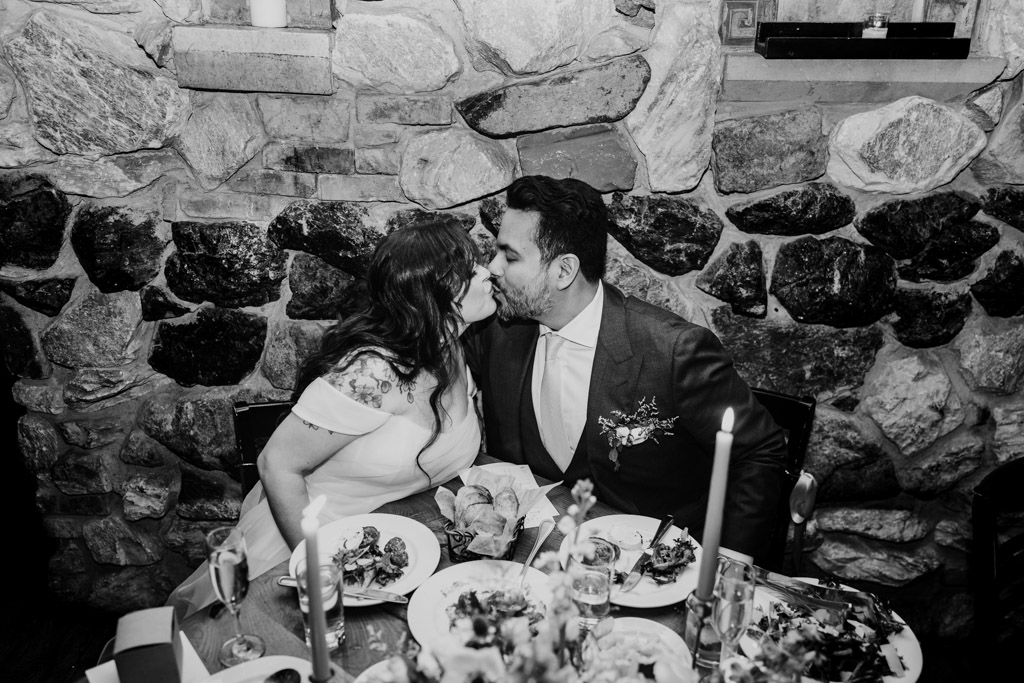 Bride and groom kiss at a romantic, candlelit dinner table set with food and drinks at Osteria Via Stato
