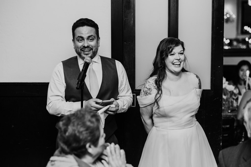 A man speaks at a microphone while a woman in a wedding dress stands smiling beside him at Osteria Via Stato.