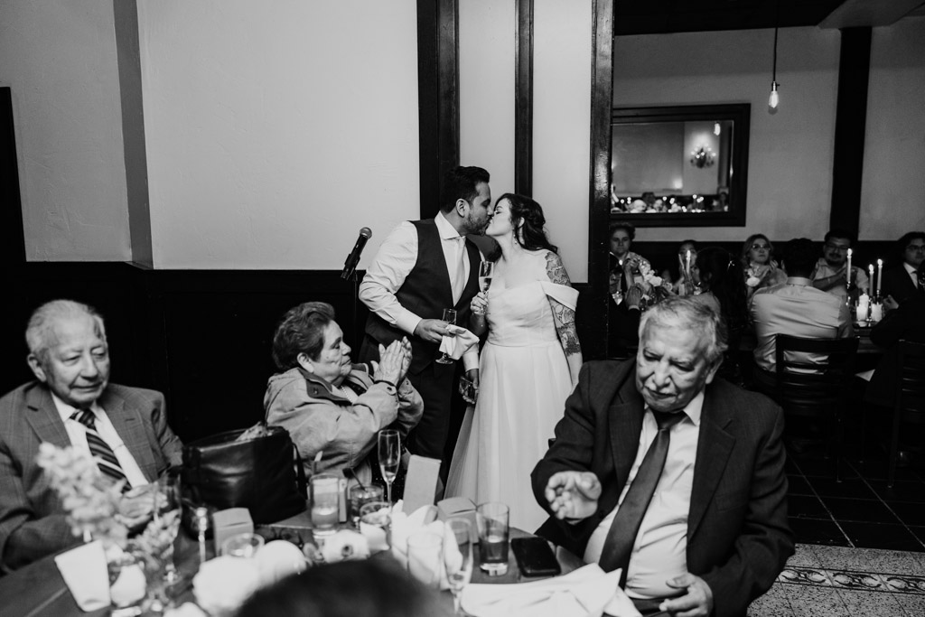Black and white photo of bride and groom kissing while guests seated at tables watch and celebrate during their Osteria Via Stato wedding reception