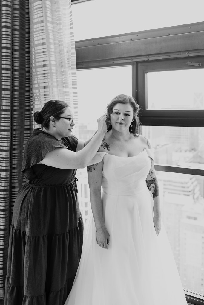 Black and white photo of bridesmaid helps bride with her hair by a window overlooking downtown Chicago buildings before her Osteria Via Stato wedding celebration