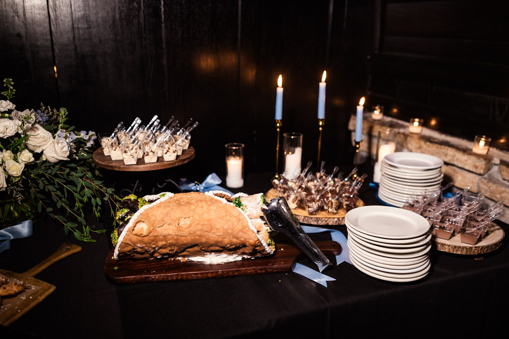 Dessert table featuring arge cannoli, mini desserts, plates, and candles on a black tablecloth for wedding reception at Osteria Via Stato