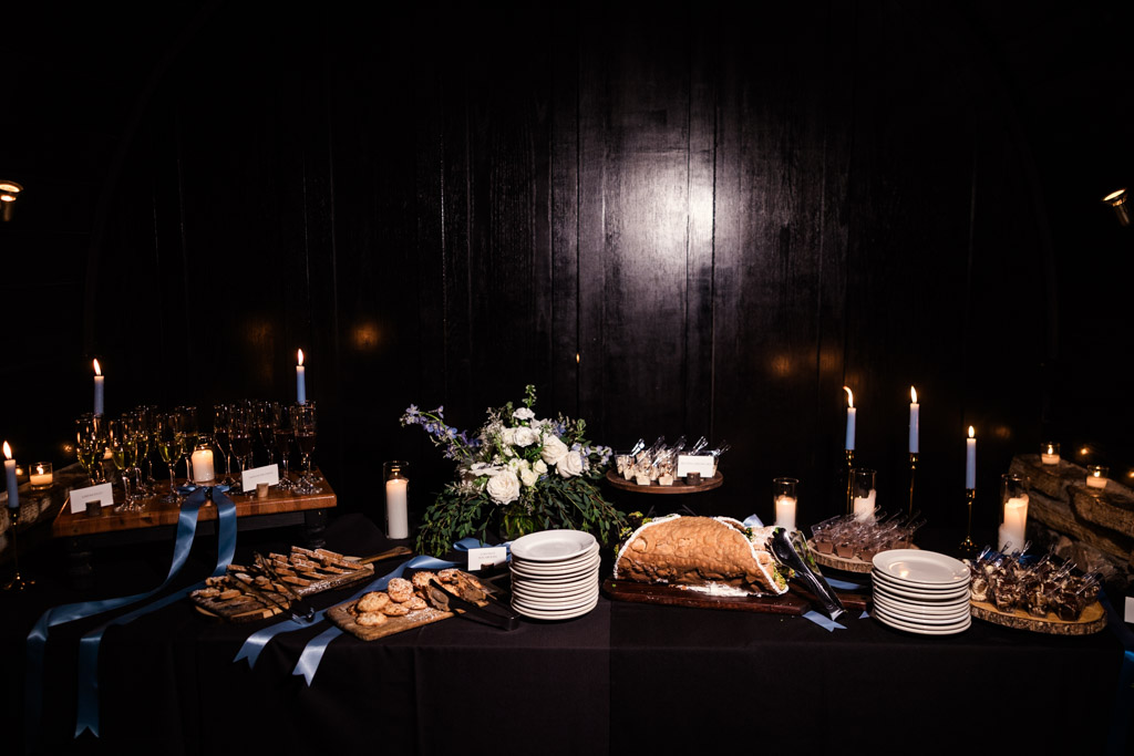 A candlelit dessert table at Osteria Via Stato features plates, pastries, cookies, a floral arrangement, and blue ribbon decorations