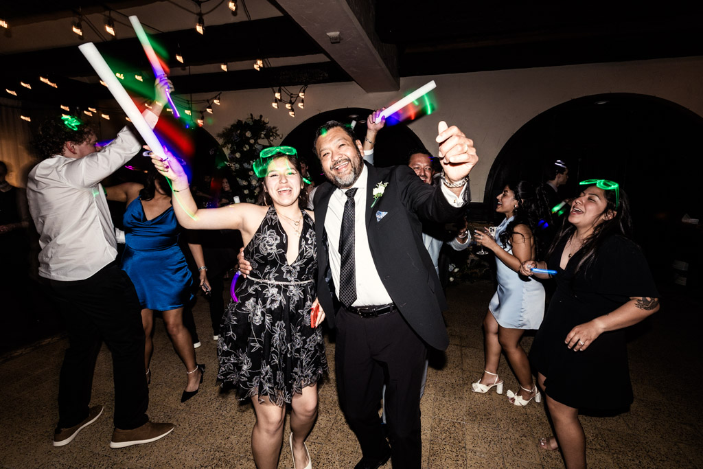People in formal wear dance and celebrate with glow sticks at a lively Osteria Via Stato party.