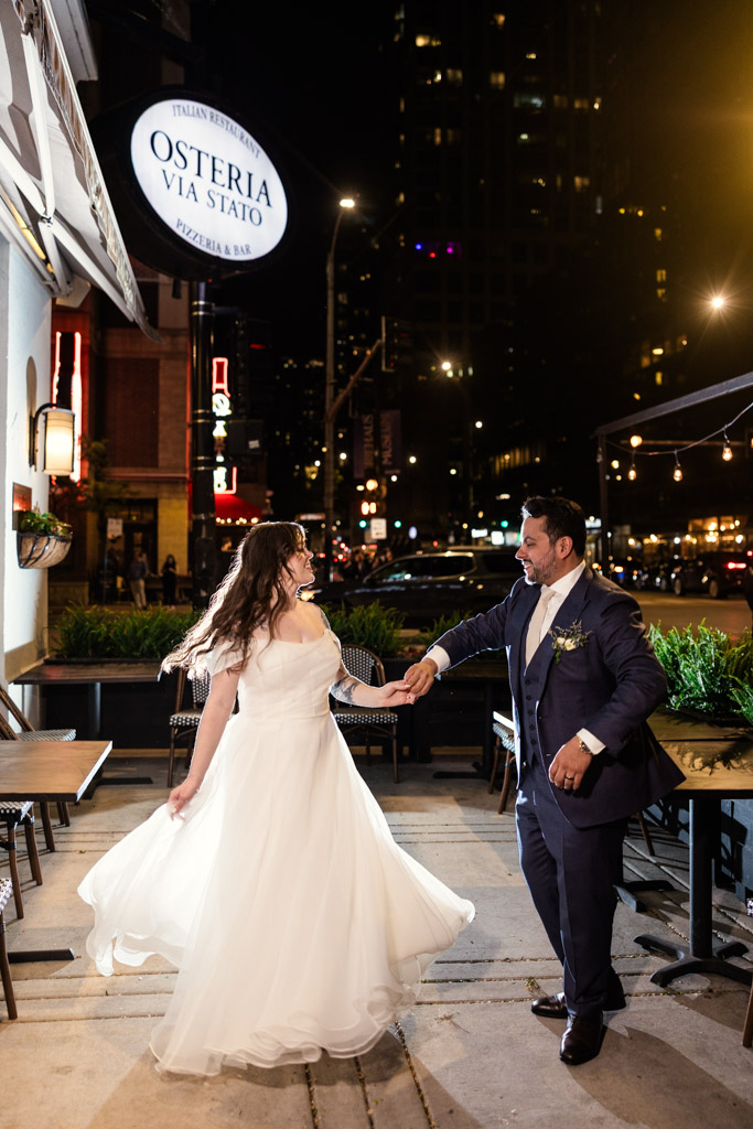 A bride and groom dance joyfully outside Osteria Via Stato at night on a city sidewalk
