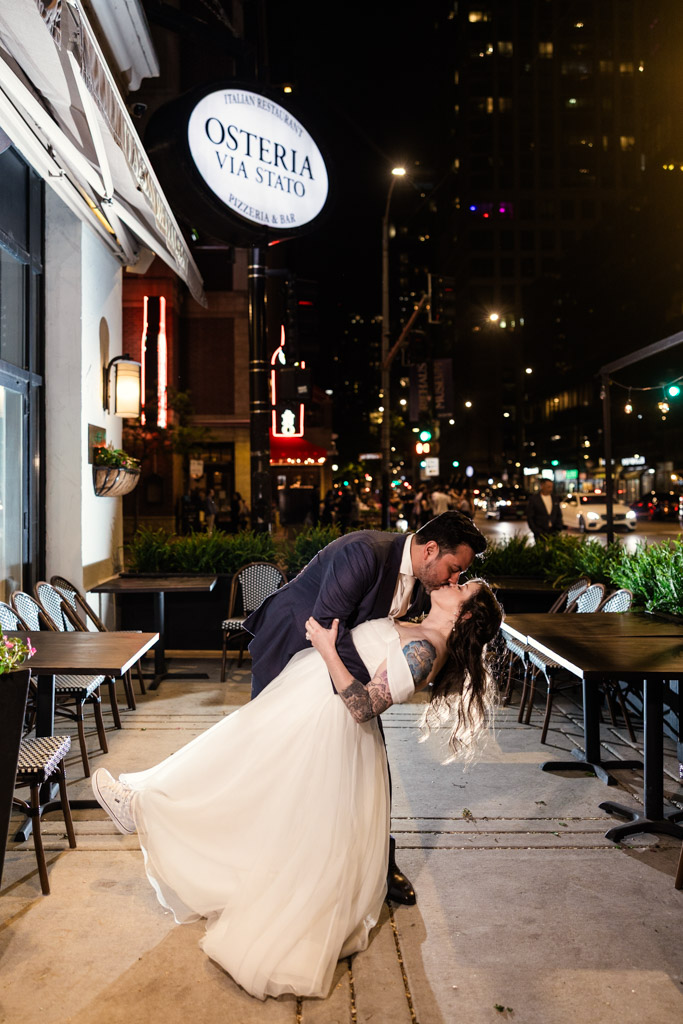 Newlyweds in wedding attire kiss under the city lights outside Osteria Via Stato