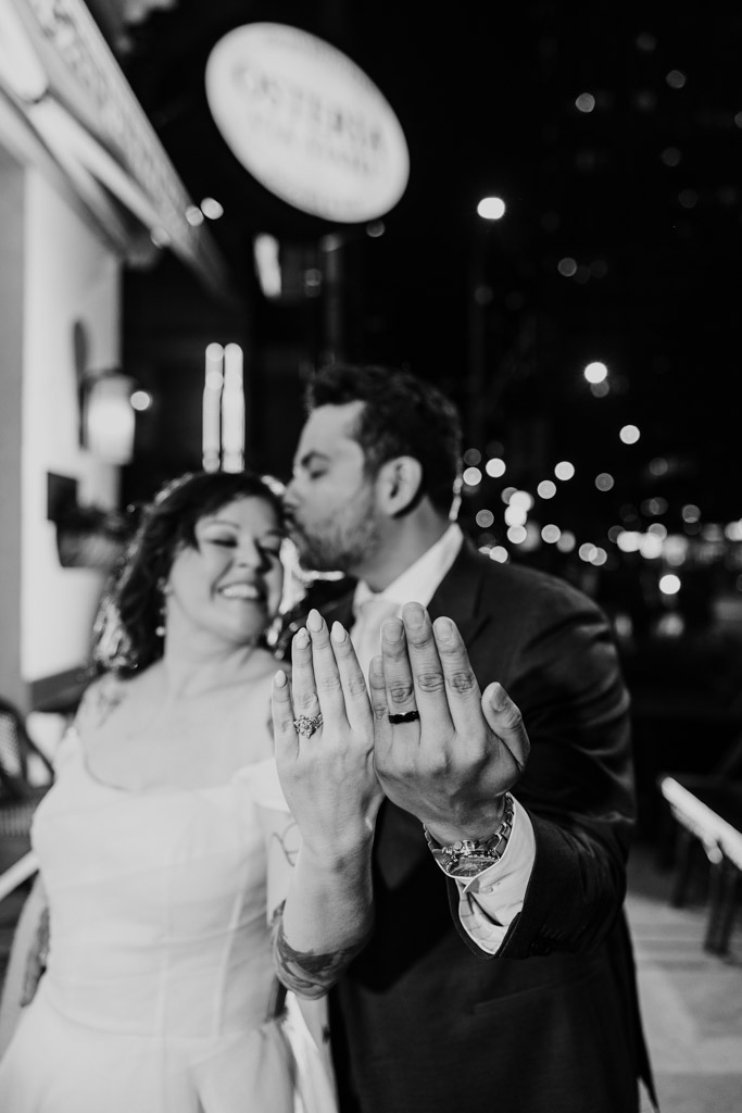 Black and white photo of bride and groom showing their wedding rings outside Osteria Via Stato, as the groom kisses the bride’s forehead at night