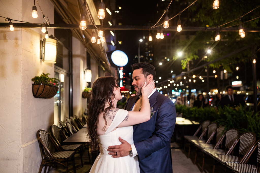 A bride and groom embrace outdoors at night under string lights outside Osteria Via Stato