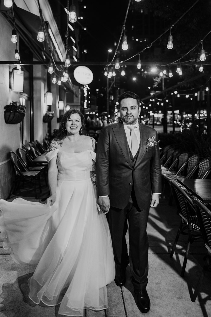 Black and white photo of bride and groom walking hand in hand at night under string lights outside Osteria Via Stato in downtown Chicago