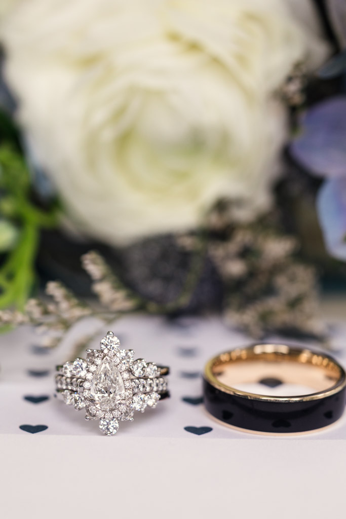 Diamond engagement ring and black wedding band on a surface with small heart shapes, flowers in the background for wedding celebration at Osteria Via Stato