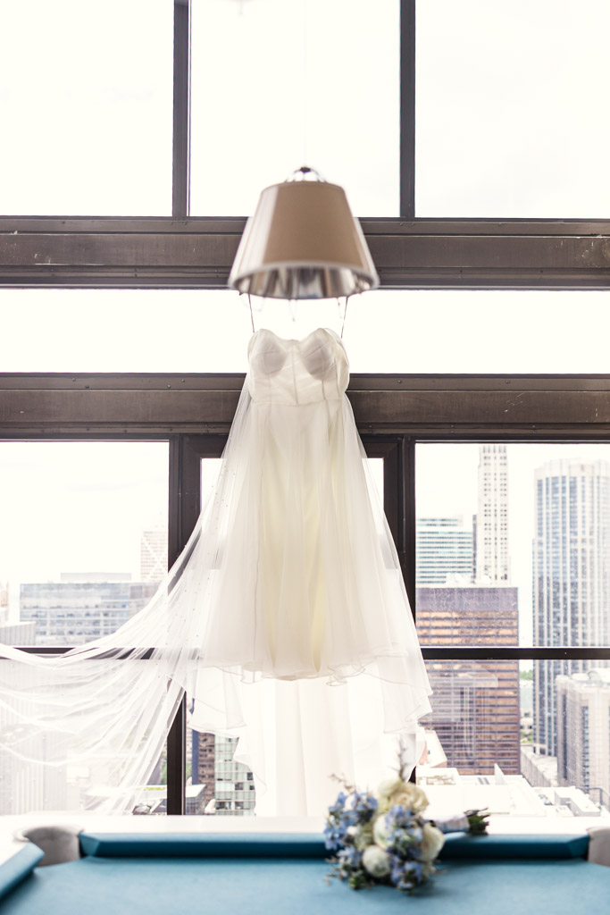 A white wedding dress hangs in front of large windows overlooking the Chicago skyline, with a bouquet below forwedding celebration at Osteria Via Stato