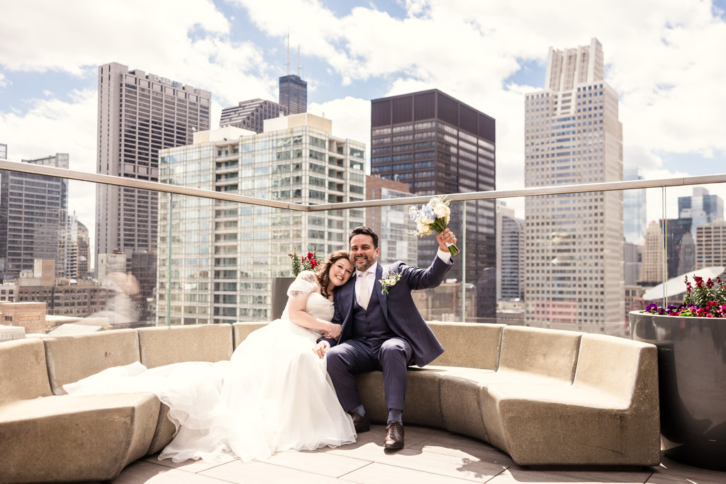 Bride and groom sit and smile on the rooftop patio of Cerise in Virgin Hotels Chicago, with city skyscrapers in the background