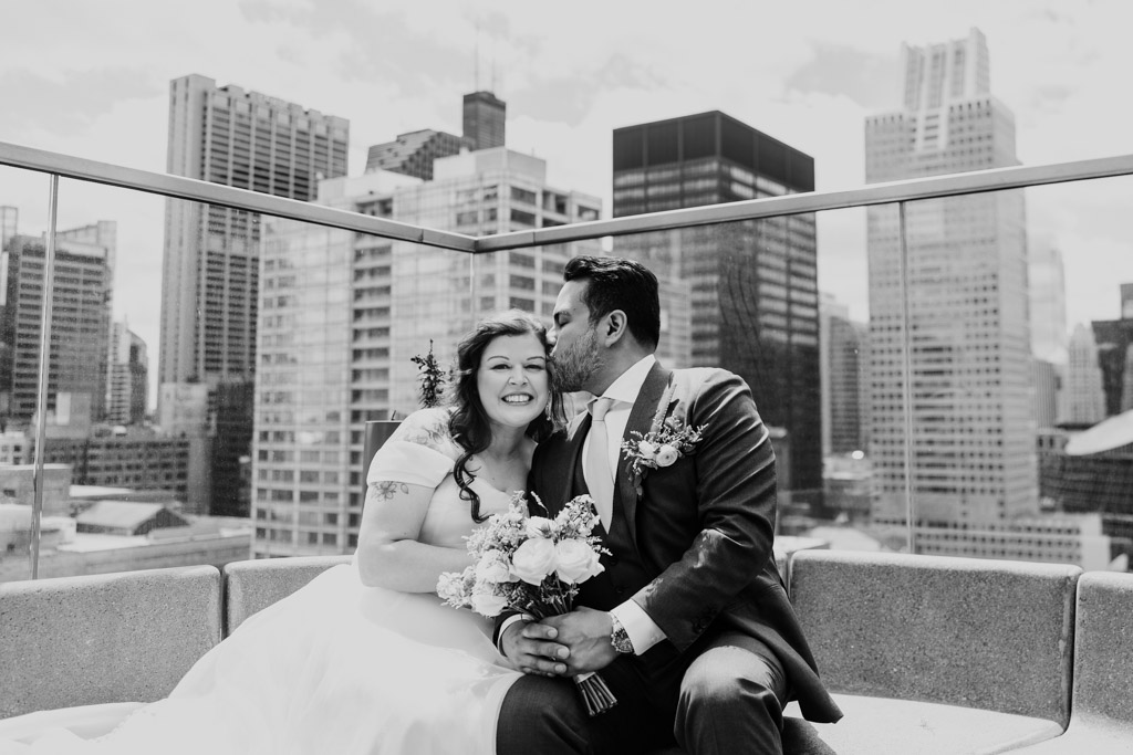Black and white photo of groom kissing bride’s cheek as they sit on a rooftop sofa at Cerise in Virgin Hotels Chicago