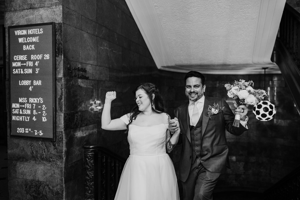Black and white photo of bride and groom smiling and celebrating while walking up stairs at Virgin Hotels Chicago, holding hands and flowers