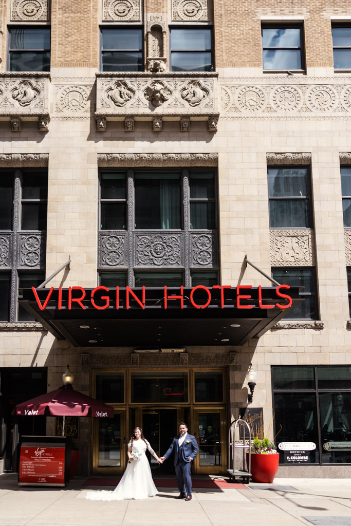 A bride and groom hold hands outside the entrance of Virgin Hotels Chicago on a sunny day