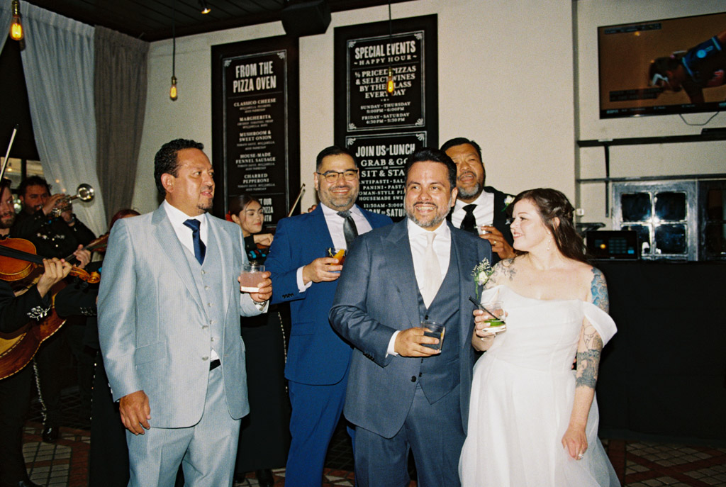 A bride and four men in suits smile and hold drinks at an Osteria Via Stato wedding reception with a mariachi band.