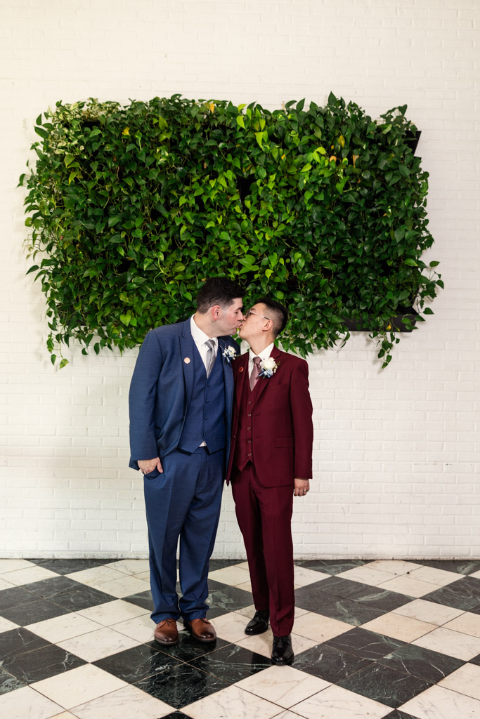 Two grooms in suits share a kiss in front of a green plant wall and white brick background at The Drake hotel in Oak Brook