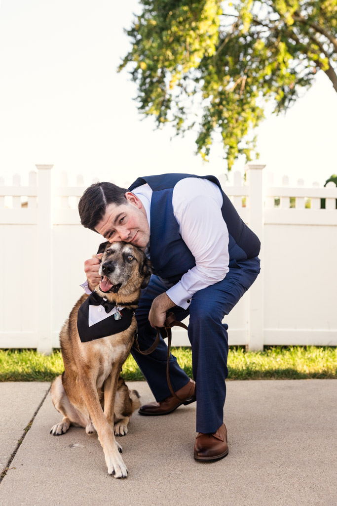 Groom in a blue suit hugs his dog by a white fence outside their home on a sunny day