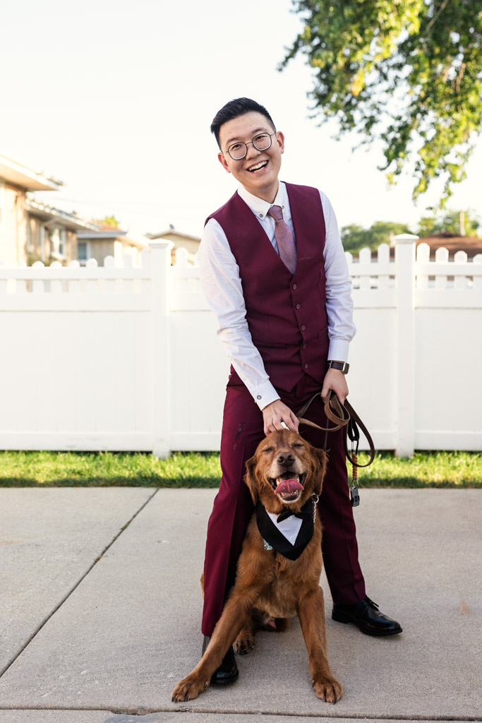 A smiling groom in a burgundy suit stands outside with a happy dog wearing a black bow tie near his home
