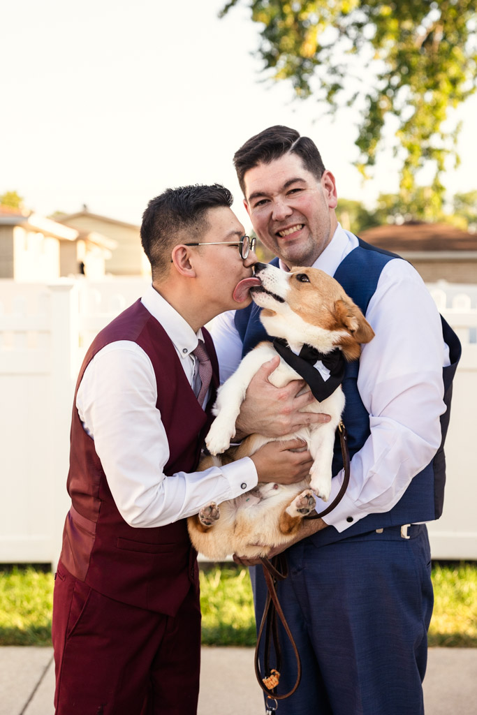 Two grooms smiling outside their home as a corgi in a bow tie licks one man's face on a sunny day