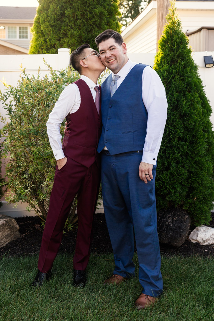 Groom in maroon suit kisses his partner wearing a blue suit on the cheek outdoors at The Drake Hotel