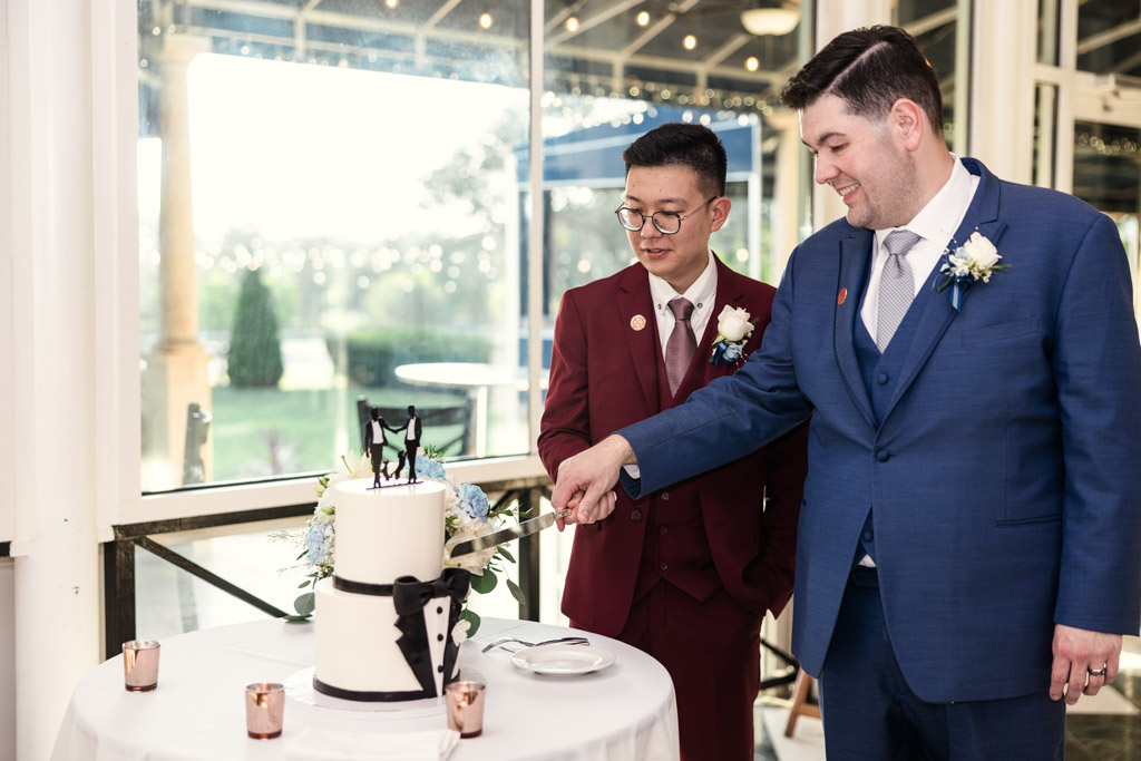 Two grooms in suits cut a white wedding cake with a tuxedo design and figurine topper at The Drake Hotel in Oak Brook