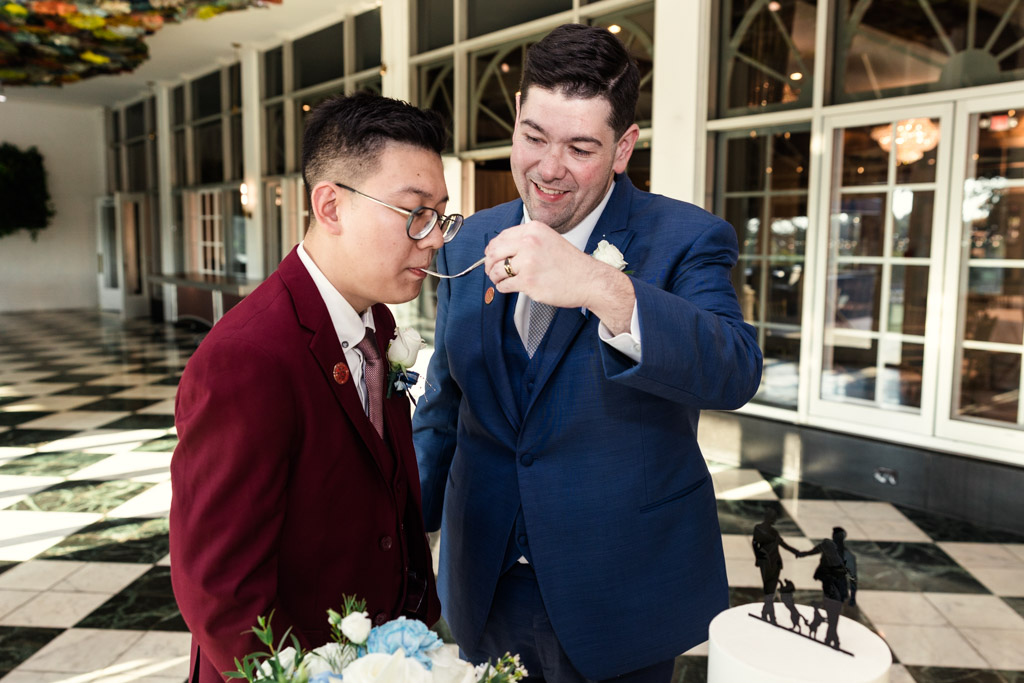 Two grooms in suits share a piece of cake at The Drake Oak Brook wedding, standing beside a white cake with a decorative topper