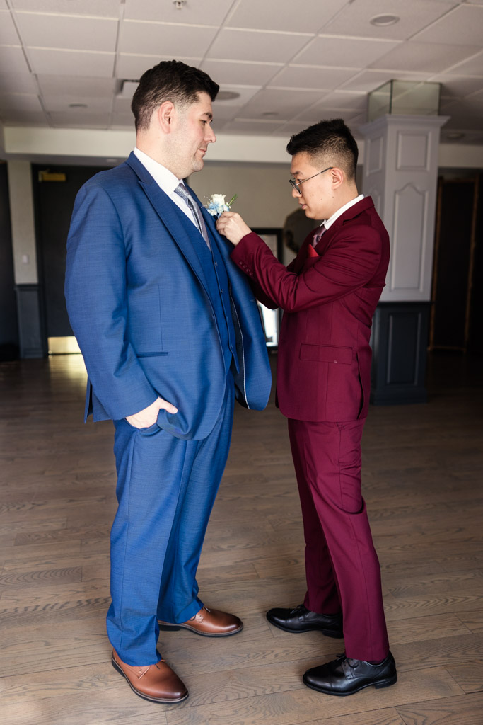 Groom in a burgundy suit adjusts his partner's boutonniere as they get ready for their wedding ceremony at The Drake Oak Brook