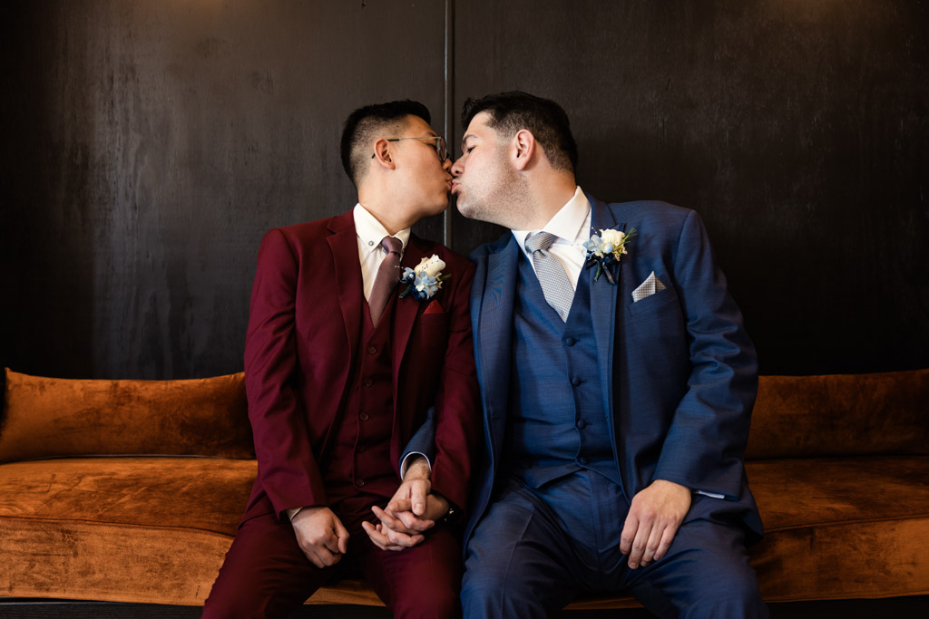 Two grooms in suits kiss while sitting on a brown couch at The Drake Oak Brook, holding hands and wearing boutonnieres