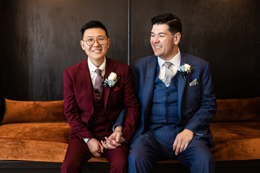 Two grooms in suits sit on a couch at The Drake Oak Brook, holding hands and smiling, with boutonnières on their jackets