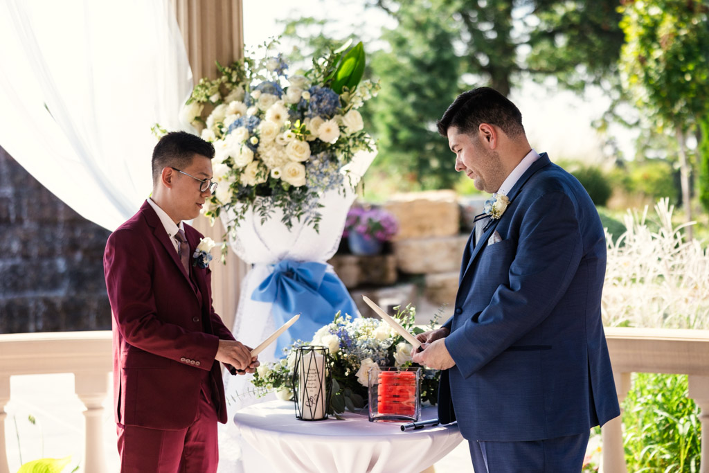 Two grooms in suits light a unity candle, during their outdoor wedding ceremony at The Drake Oak Brook