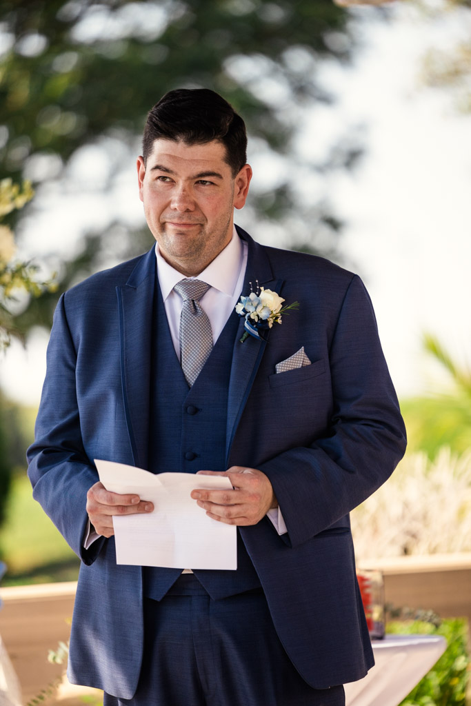 Groom in a blue suit holding his vows, standing outdoors with a boutonniere on his jacket during wedding ceremony at The Drake