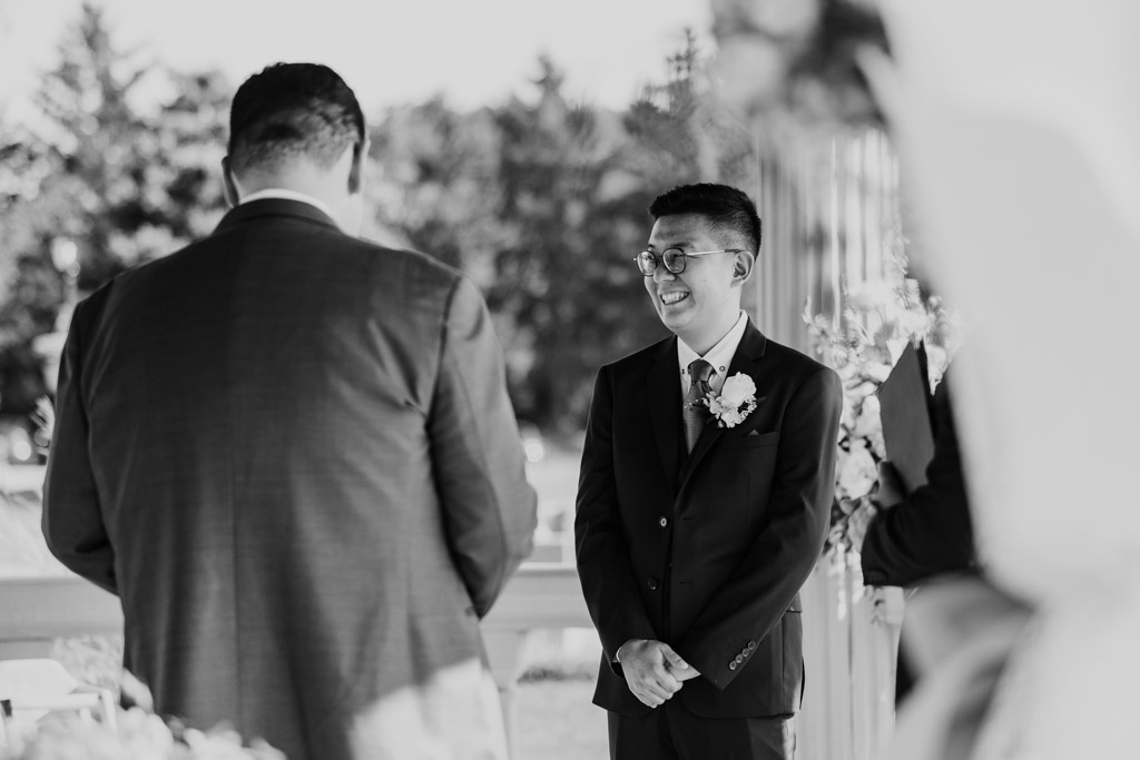 Black and white photo of groom in a suit smiling at his partner during their outdoor wedding ceremony at The Drake in Oak Brook, Illinois