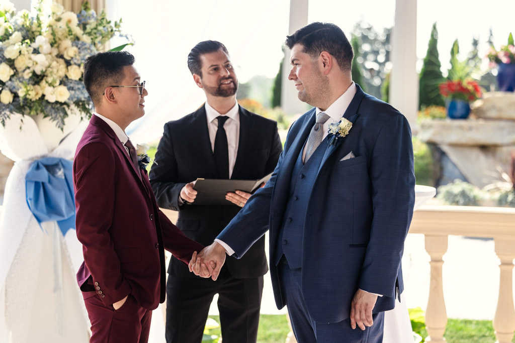 Two grooms hold hands and smile at each other during their outdoor elopement ceremony with an officiant present at The Drake in Oak Brook
