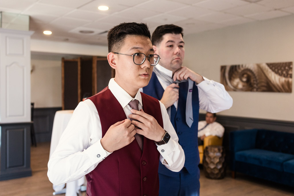 Grooms in suits adjust their ties in a bright room at The Drake, preparing for their outdoor wedding ceremony