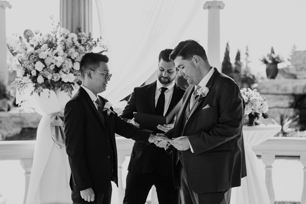 Black adn white photo of two grooms exchanging rings during an outdoor wedding ceremony at The Drake, with the officiant standing behind them