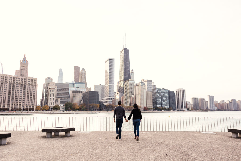 A couple holds hands while walking toward the Chicago skyline along a waterfront promenade in Olive Park, during their Chicago fall engagement session