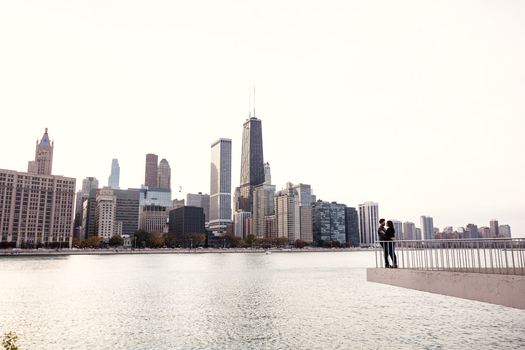 A couple embraces on a pier in Olive Park with the Chicago skyline in the background across the water during fall engagement session
