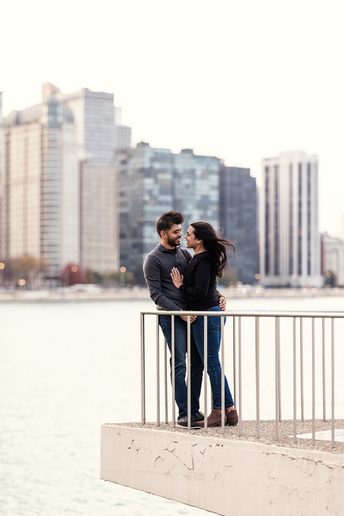 A couple stands close together by a railing at Olive Park, with city buildings and Lake Michigan in the background during Chicago fall engagement session