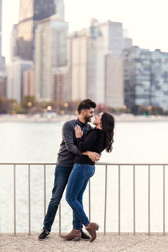 Engaged couple embraces and smiles at each other on the lakefront, with Chicago’s skyline in the background