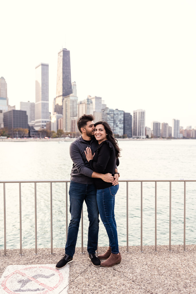 Engaged couple embraces by a waterfront railing in Olive Park with the Chicago skyline in the background