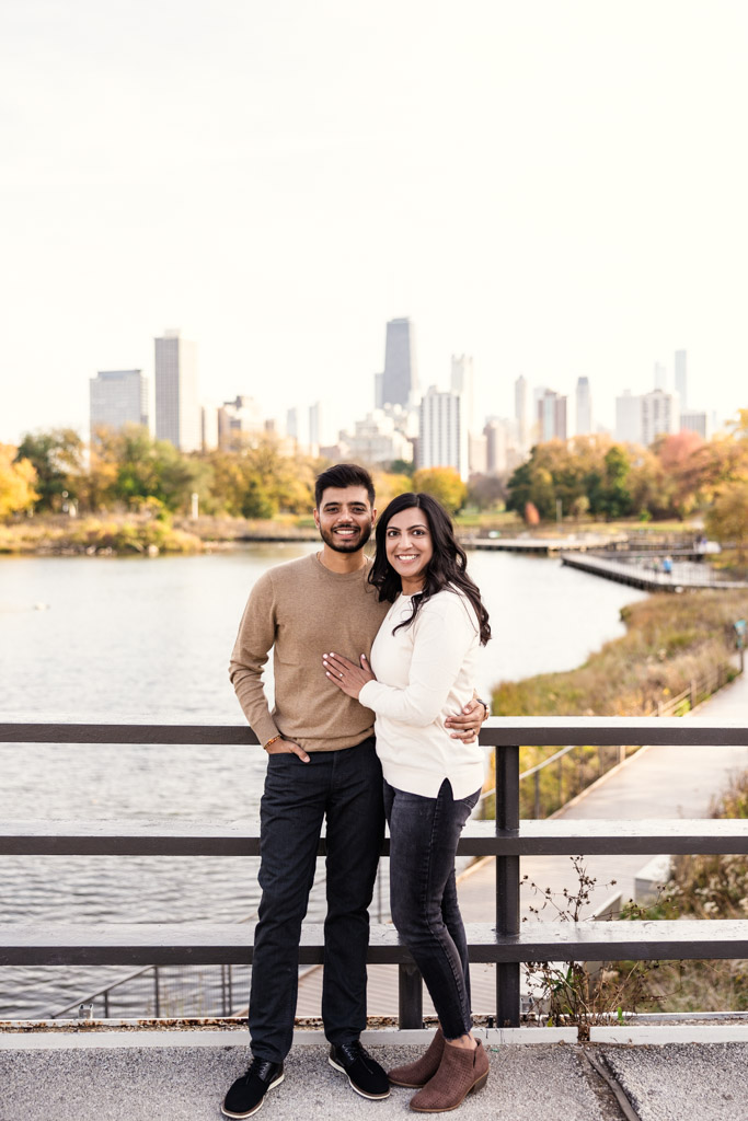 Portrait of happy engaged couple stands by a Lincoln Park bridge railing with Chicago skyline and pond in the background