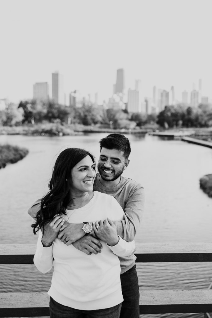 Black and white photo of happy engaged couple embracing on a Lincoln Park bridge with the Chicago skyline in the background