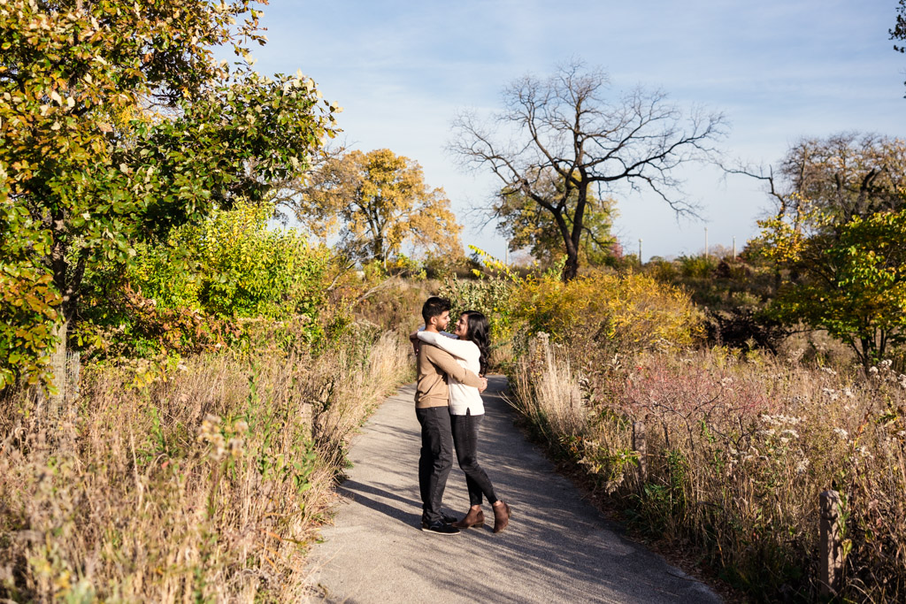 A couple embraces on a sunny Lincoln Park path, surrounded by autumn trees and tall grasses during their Chicago fall engagement session