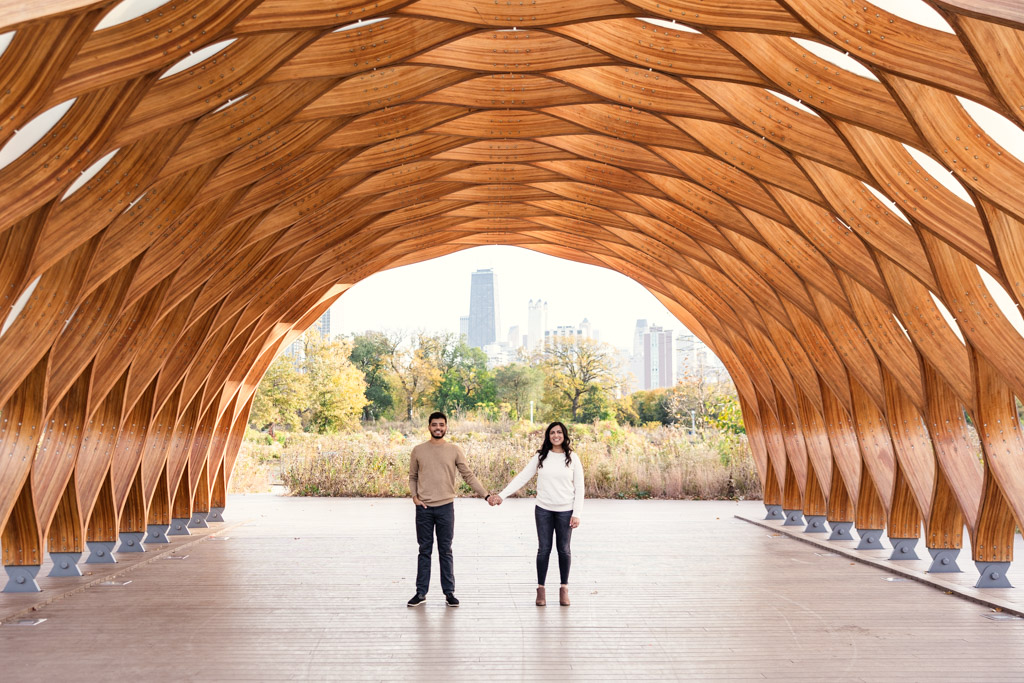 Portrait of engaged couple holding hands under the Lincoln Park Honeycomb with the Chicago skyline in the background