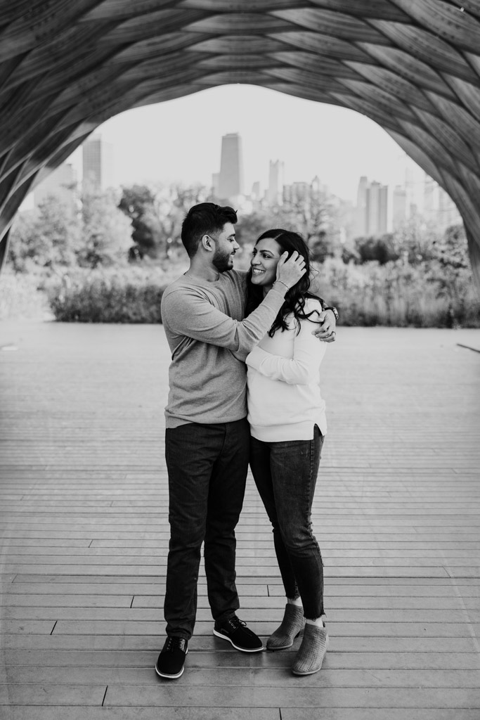Black and white photo of couple standing smiling under the Lincoln Park Honeycomb with the Chicago skyline in the background during their fall engagement session